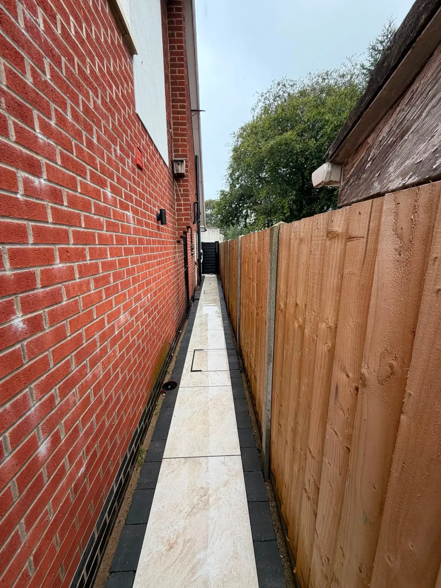 A narrow paved side path between a brick wall and a timber fence, laid with matching stone-effect paving, still wet after cleaning — from the same completed job by Midland Revive.
