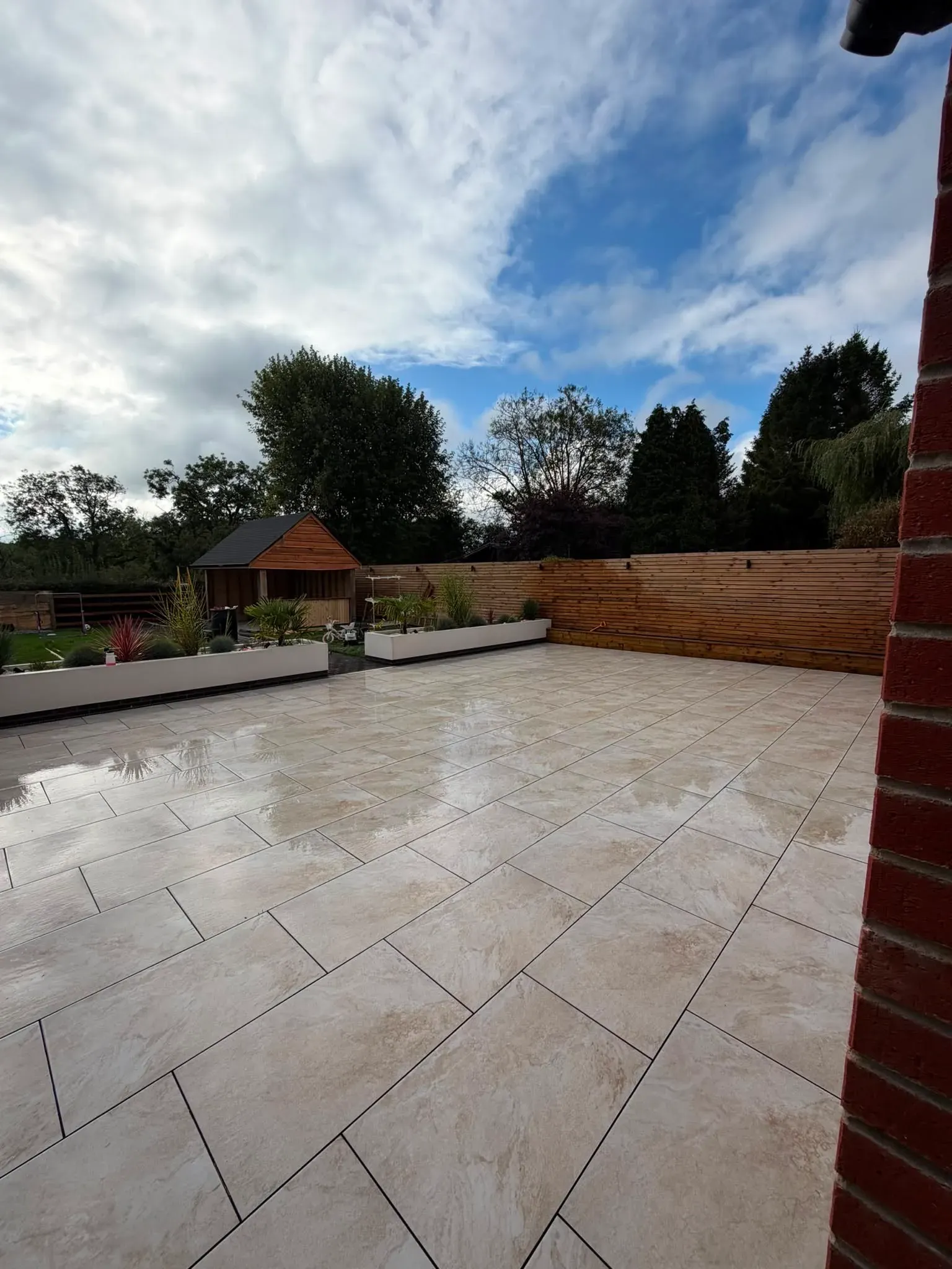 A large patio area laid with light stone-effect paving, still wet after cleaning, bordered by raised planters and a timber fence — a completed patio cleaning job by Midland Revive.