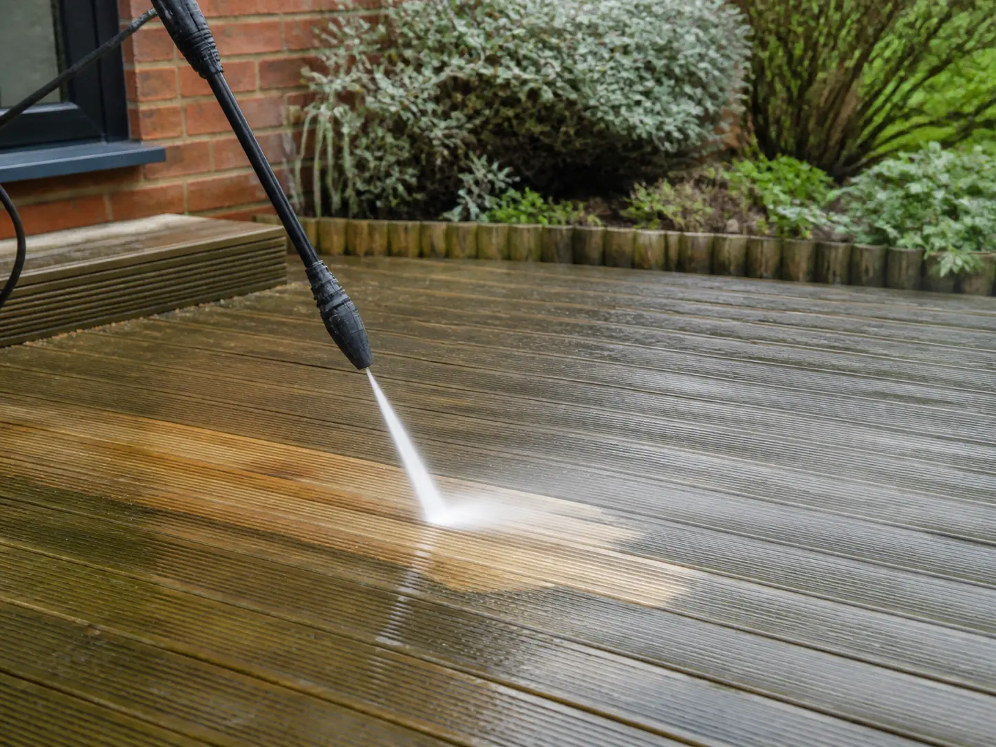 Weathered timber garden decking being cleaned with a controlled pressure-washer lance.